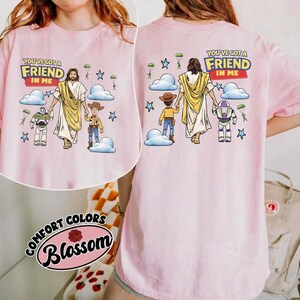 May include: Pale pink t-shirt with a graphic of Jesus, Woody, and Buzz Lightyear, with the text "YOU'VE GOT A FRIEND IN ME." The design includes clouds, stars, and parachutes. The brand name "Comfort Colors Blossom" is printed on the bottom.