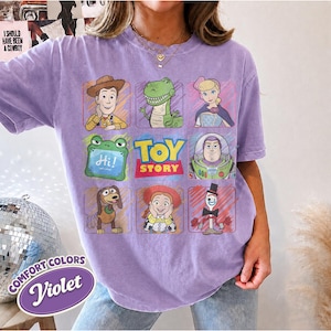 May include: A light purple short-sleeve t-shirt featuring a grid of nine Toy Story character illustrations including Woody, Rex, Bo Peep, Slinky Dog, Jessie, and Forky, with the Toy Story logo in the center and a green frog holding a sign saying Hi.
