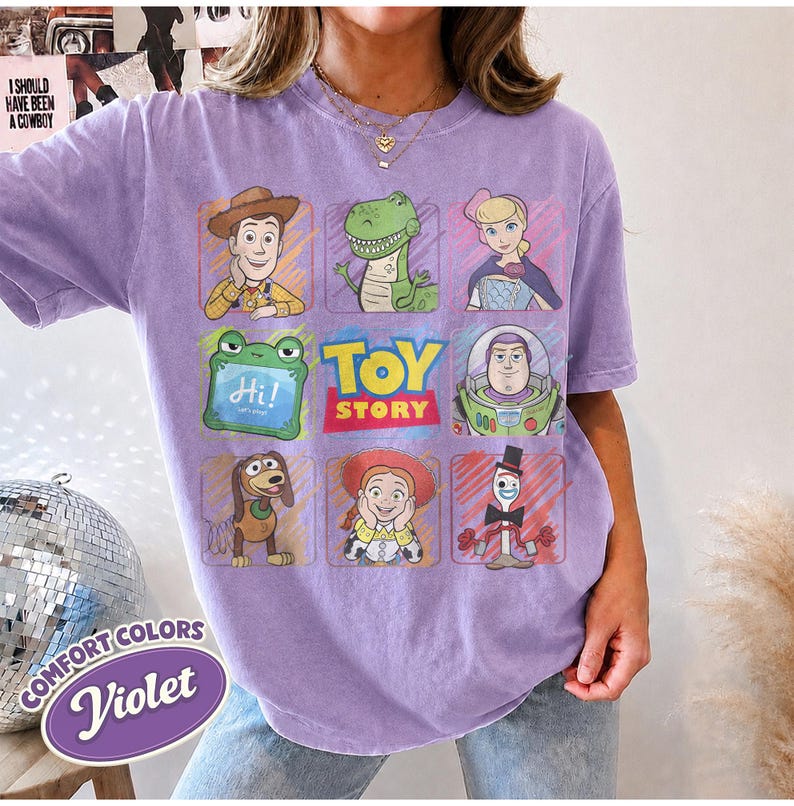 May include: A light purple short-sleeve t-shirt featuring a grid of nine Toy Story character illustrations including Woody, Rex, Bo Peep, Slinky Dog, Jessie, and Forky, with the Toy Story logo in the center and a green frog holding a sign saying Hi.