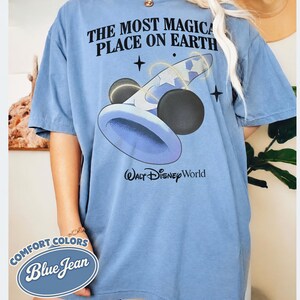 K&ouml;nnte beinhalten: Blaues Jeans-T-Shirt mit dem Text "THE MOST MAGICAL PLACE ON EARTH" und einer Grafik eines Zauberhuts und Mickey-Mouse-&auml;hnlichen Kugeln. Die Worte "Walt Disney World" sind darunter gedruckt. Das Shirt hat auch ein "Comfort Colors Blue Jean"-Etikett.