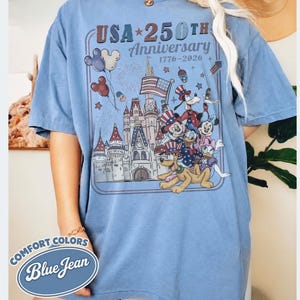 Puede incluir: Camiseta azul claro con un dise&ntilde;o gr&aacute;fico que celebra el 250 aniversario de EE. UU., con personajes de Disney, un castillo y fuegos artificiales. El dise&ntilde;o incluye el texto "USA 250th Anniversary 1776-2026" y las palabras "Comfort Colors Blue Jean".
