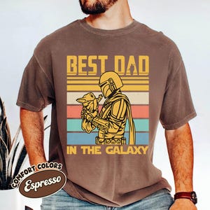 以下が含まれることがあります： エスプレッソ色のTシャツ。マンダロリアン風のキャラクターが小さな生き物を抱いているグラフィックが特徴です。「BEST DAD IN THE GALAXY」の文字が黄色で描かれています。ヴィンテージ感のあるシャツです。