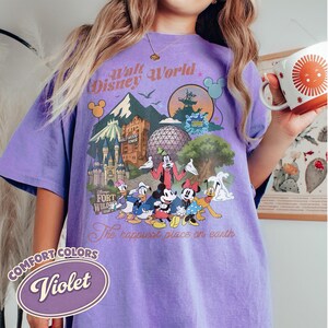 Pu&ograve; includere: Una maglietta Comfort Colors viola con una grafica colorata dei personaggi e dei monumenti di Walt Disney World. Il design include Topolino, Minnie, Pippo e Paperino, con il testo "Walt Disney World" e "The happiest place on earth."