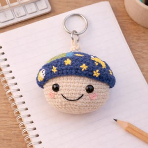 May include: A crocheted keychain shaped like a smiling mushroom with a blue cap adorned with yellow stars. The mushroom has a cream-colored body, rosy cheeks, and a silver keyring.