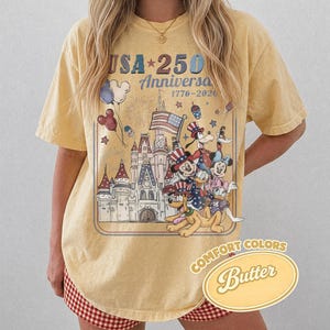 Puede incluir: Camiseta amarilla con un gr&aacute;fico de personajes de Disney celebrando el 250 aniversario de EE. UU. El dise&ntilde;o incluye un castillo, fuegos artificiales y la bandera estadounidense. La camiseta tiene el texto "USA 250 Anniversa 1776-2026" y "Comfort Colors Butter".