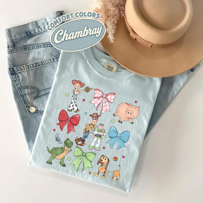 May include: A light blue t-shirt featuring a graphic print of various cartoon characters and colorful bows. The shirt is displayed alongside a pair of light wash denim jeans, a tan wide-brimmed hat, and a silver necklace. Text on the shirt reads Comfort Colors Chambray.