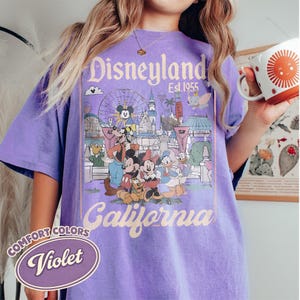 Może przedstawiać: Fioletowa koszulka Comfort Colors z grafiką Disneyland California. Projekt zawiera Myszkę Miki, Minnie, Kaczora Donalda i inne postacie Disneya przed sceną parku. Koszulka ma wygląd vintage.