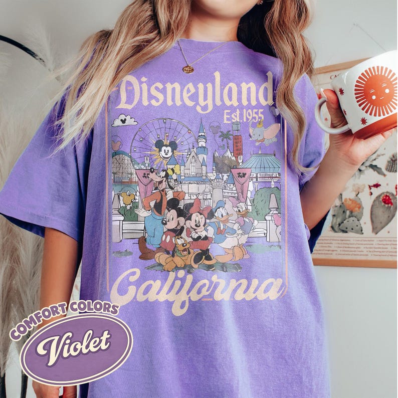 Może przedstawiać: Fioletowa koszulka Comfort Colors z grafiką Disneyland California. Projekt zawiera Myszkę Miki, Minnie, Kaczora Donalda i inne postacie Disneya przed sceną parku. Koszulka ma wygląd vintage.