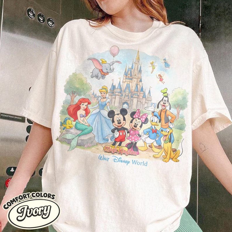Op de afbeelding: Een ivoorkleurig t-shirt met een kleurrijke grafische print van diverse stripfiguren waaronder Mickey Mouse, Minnie Mouse, Donald Duck, Goofy, Pluto, Ari&euml;l en Dumbo voor een kasteel. De tekst luidt Magic Kingdom Walt Disney World.