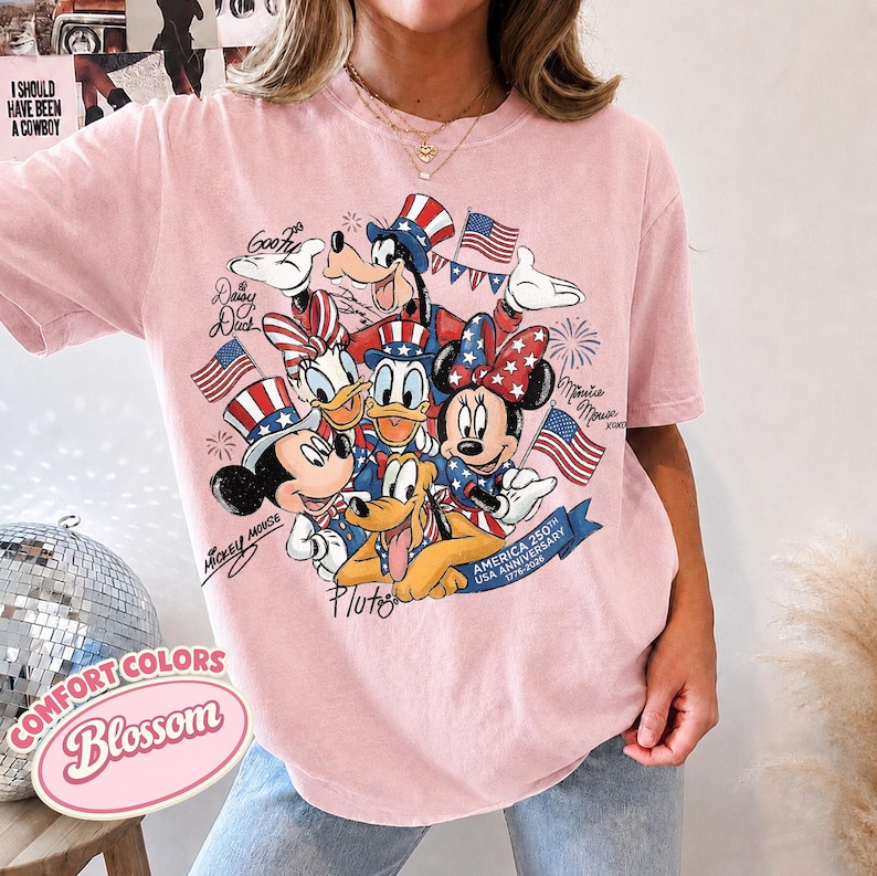 May include: Pink t-shirt featuring a graphic of Mickey Mouse, Minnie Mouse, Donald Duck, Daisy Duck, Goofy, and Pluto in patriotic attire. The design includes American flags and text that reads "America 250th USA Anniversary 1776-2026". The shirt also has the text "Comfort Colors Blossom".