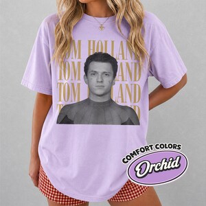 Peut inclure: T-shirt lilas avec une image en noir et blanc de Tom Holland en Spider-Man. Les mots "TOM HOLLAND" sont imprim&eacute;s en or. Le t-shirt porte &eacute;galement une &eacute;tiquette Comfort Colors Orchid.