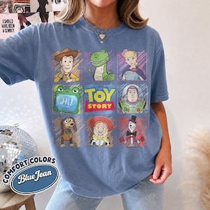 May include: A blue jean colored t-shirt featuring a grid graphic of Toy Story characters including Woody, Rex, Bo Peep, a green frog, Buzz Lightyear, Slinky Dog, Jessie, and Forky, with the Toy Story logo centered in the middle of the design.