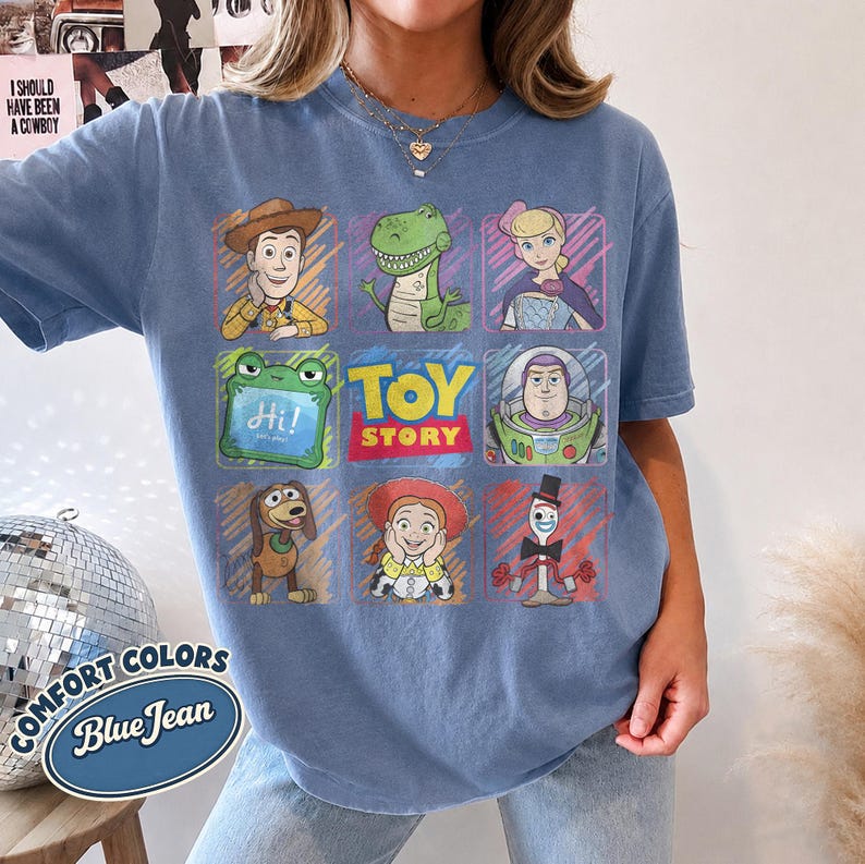 May include: A blue jean colored t-shirt featuring a grid graphic of Toy Story characters including Woody, Rex, Bo Peep, a green frog, Buzz Lightyear, Slinky Dog, Jessie, and Forky, with the Toy Story logo centered in the middle of the design.