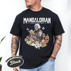 May include: Black t-shirt featuring a graphic of The Mandalorian and Grogu surrounded by flowers and butterflies. The text "MANDALORIAN - THIS IS THE WAY" is printed across the top. The shirt has a Comfort Colors label.