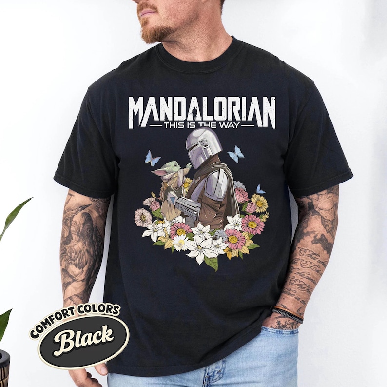 May include: Black t-shirt featuring a graphic of The Mandalorian and Grogu surrounded by flowers and butterflies. The text "MANDALORIAN - THIS IS THE WAY" is printed across the top. The shirt has a Comfort Colors label.