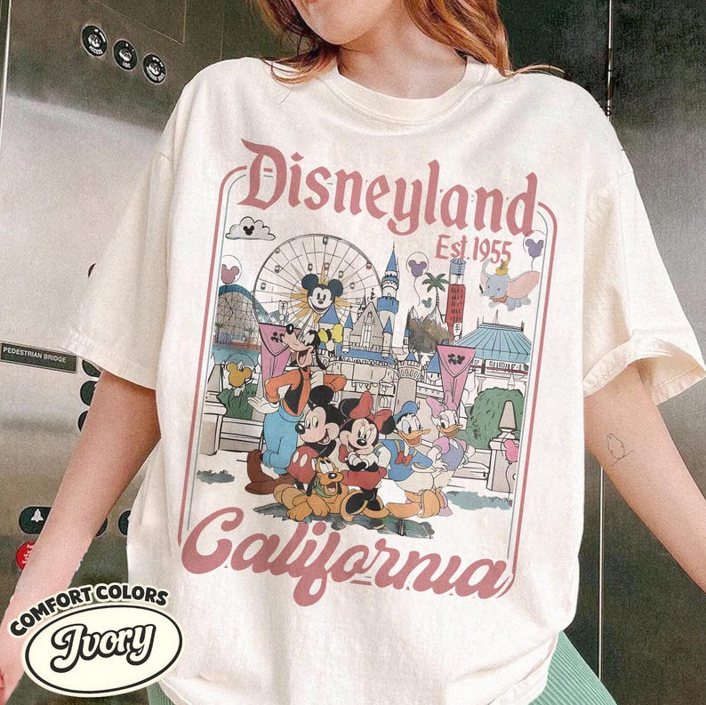 Może przedstawiać: Kremowa koszulka z grafiką Disneyland. Projekt przedstawia Myszka Miki, Minnie, Kaczora Donalda i inne postacie Disneya przed diabelskim młynem i zamkiem. Tekst "Disneyland Est. 1955 California" jest wydrukowany w r&oacute;żowo-czerwonym kolorze.