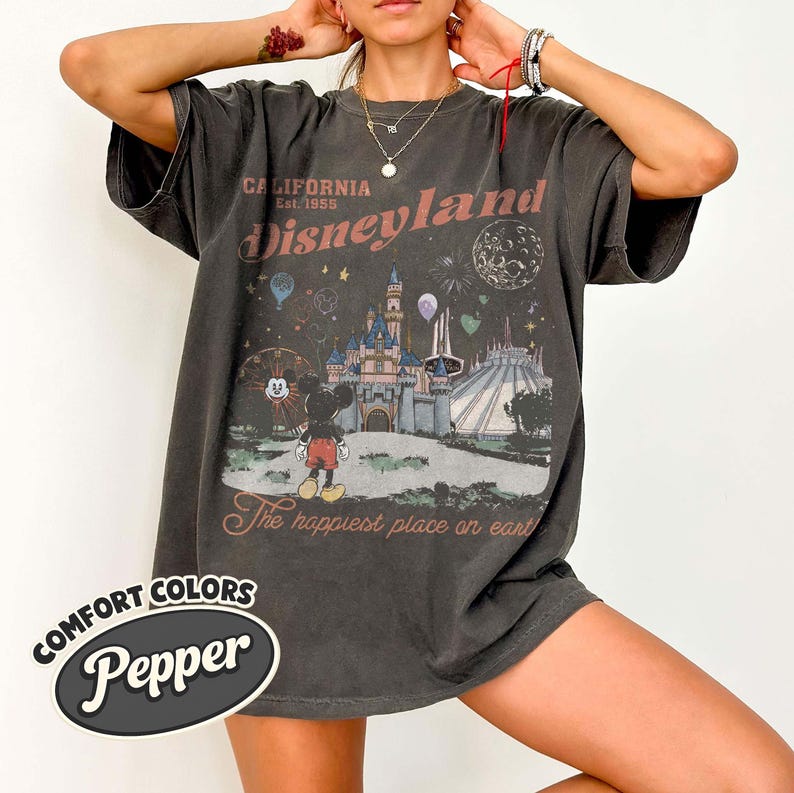 May include: Dark gray t-shirt with a Disneyland graphic. The design features a castle, Mickey Mouse, fireworks, and the text "California Est. 1955 Disneyland" and "The happiest place on earth." The shirt has a Comfort Colors Pepper label.