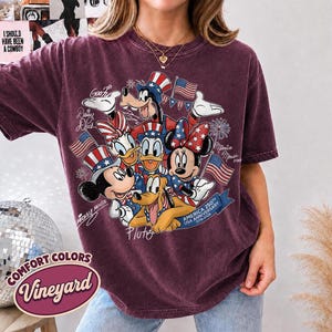 May include: A maroon Comfort Colors t-shirt featuring a graphic of Disney characters Mickey Mouse, Minnie Mouse, Donald Duck, Daisy Duck, Goofy, and Pluto, celebrating the USA's 250th anniversary. The design includes American flags and patriotic hats.