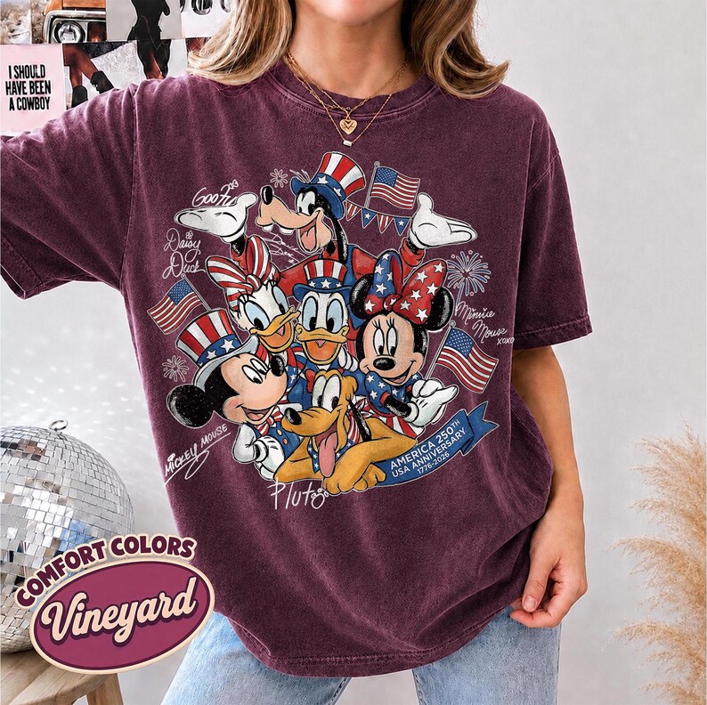 May include: A maroon Comfort Colors t-shirt featuring a graphic of Disney characters Mickey Mouse, Minnie Mouse, Donald Duck, Daisy Duck, Goofy, and Pluto, celebrating the USA's 250th anniversary. The design includes American flags and patriotic hats.