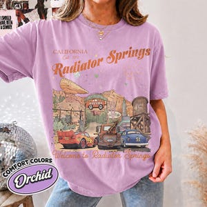 May include: A light purple t-shirt featuring a graphic print of the Radiator Springs landscape with the text California Est. 2017, Radiator Springs, and Welcome to Radiator Springs. The design includes a red race car, a brown tow truck, and a blue car.