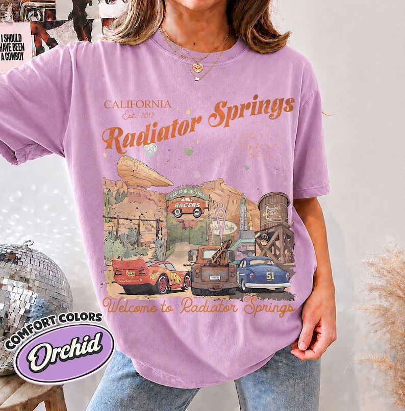 May include: A light purple t-shirt featuring a graphic print of the Radiator Springs landscape with the text California Est. 2017, Radiator Springs, and Welcome to Radiator Springs. The design includes a red race car, a brown tow truck, and a blue car.