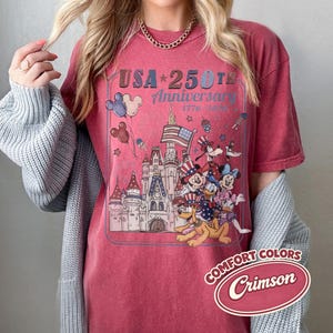 Puede incluir: Camiseta de color carmes&iacute; con un dise&ntilde;o gr&aacute;fico que celebra el 250 aniversario de EE. UU. El dise&ntilde;o presenta un castillo, Mickey Mouse, Minnie Mouse, Donald Duck y Pluto, con una bandera estadounidense y globos. La camiseta est&aacute; etiquetada como "Comfort Colors".