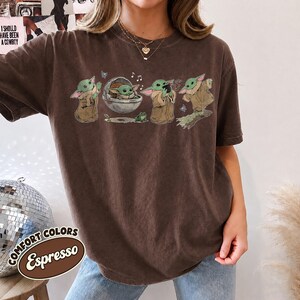 May include: A dark brown short-sleeve t-shirt featuring a graphic print of a small green creature in various poses, including sitting in a floating pod, standing, and reaching out, accompanied by small butterflies, musical notes, and a frog.