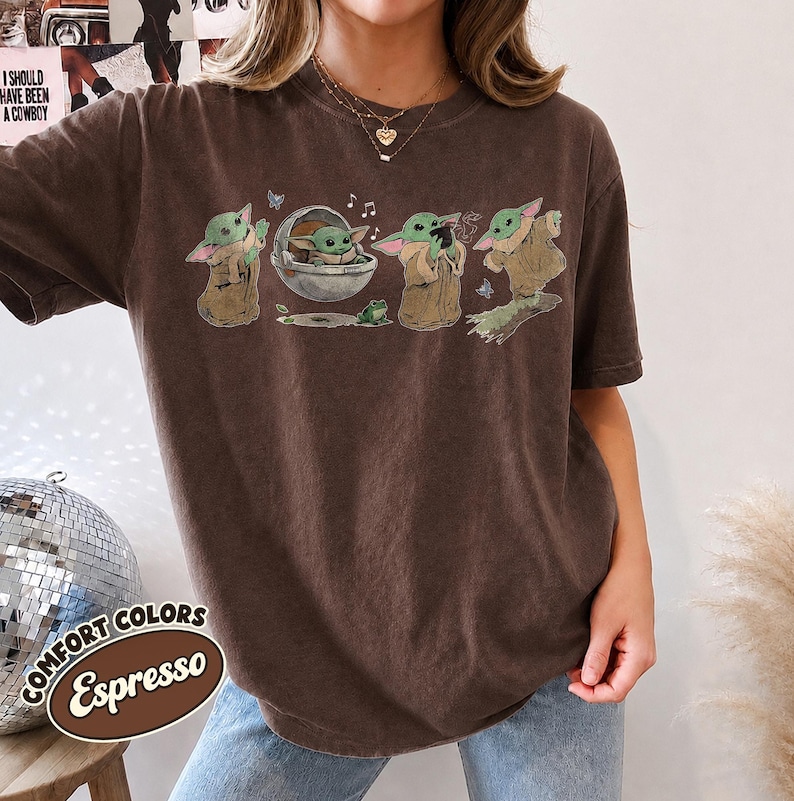 May include: A dark brown short-sleeve t-shirt featuring a graphic print of a small green creature in various poses, including sitting in a floating pod, standing, and reaching out, accompanied by small butterflies, musical notes, and a frog.