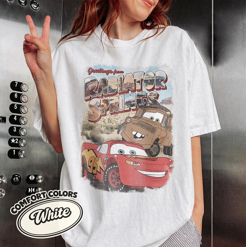 May include: White t-shirt featuring a graphic of Lightning McQueen and Mater from the movie Cars, with the text "Greetings from Radiator Springs." The shirt also has the Comfort Colors logo.