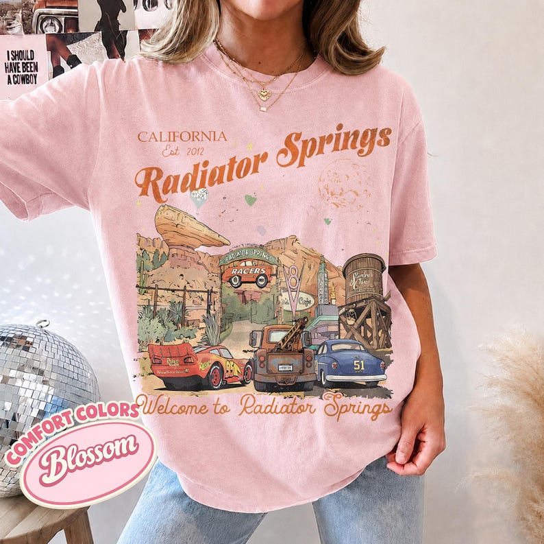 May include: A light pink short-sleeve t-shirt featuring a graphic print of the Radiator Springs landscape from the movie Cars. The design includes text that reads California Est 2012, Radiator Springs, and Welcome to Radiator Springs.