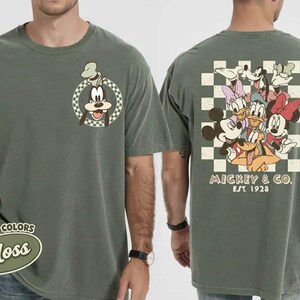 May include: Moss green t-shirt featuring a cartoon image of Goofy on the front and a group of Disney characters on the back. The back design includes Mickey Mouse, Minnie Mouse, Donald Duck, Daisy Duck, and Goofy. The text "Mickey & Co. EST 1928" is also on the back.