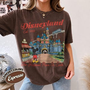 May include: A dark brown short-sleeve t-shirt featuring a graphic of the Disneyland castle, Mickey Mouse, and a monorail. The text reads Disneyland Est. 1955 and The Happiest Place On Earth in red lettering. The shirt has a crew neckline.