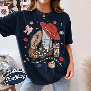 Peut inclure: Un t-shirt bleu marine fonc&eacute; avec une impression graphique d'une botte de cow-boy &agrave; motifs de vache, un chapeau rouge, un n&oelig;ud rose et des blocs empil&eacute;s avec les lettres T, S et le chiffre 13. Le texte Taylor et I KNEW IT I KNEW YOU est imprim&eacute;.