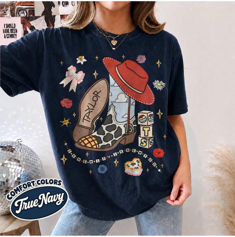 Peut inclure: Un t-shirt bleu marine fonc&eacute; avec une impression graphique d'une botte de cow-boy &agrave; motifs de vache, un chapeau rouge, un n&oelig;ud rose et des blocs empil&eacute;s avec les lettres T, S et le chiffre 13. Le texte Taylor et I KNEW IT I KNEW YOU est imprim&eacute;.
