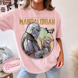 Puede incluir: Una camiseta rosa claro con un estampado gr&aacute;fico de The Mandalorian y Grogu dentro de un marco circular. El texto THE MANDALORIAN est&aacute; impreso en dorado sobre el c&iacute;rculo, con la silueta de una peque&ntilde;a nave espacial. Cuello redondo.