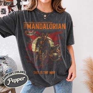 May include: A charcoal gray short-sleeve t-shirt featuring a graphic print of The Mandalorian and Grogu. The text reads "Bounty hunting is a complicated profession," "The Mandalorian," and "This is the way." The design includes a sun and red accents.