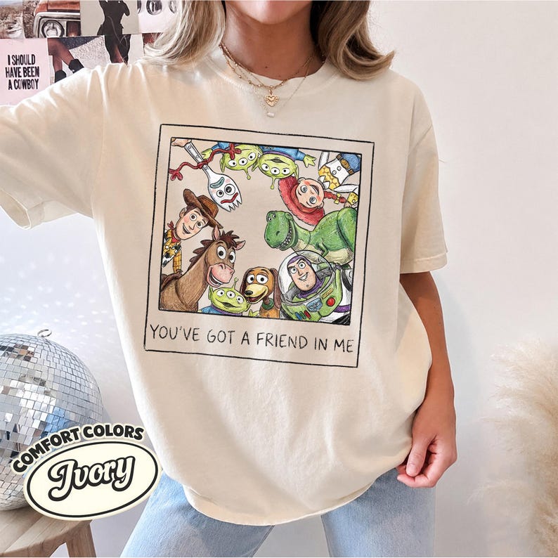 Puede incluir: Camiseta de color marfil con un gr&aacute;fico enmarcado de personajes de Toy Story. El gr&aacute;fico incluye a Woody, Buzz Lightyear y otros personajes. El texto "YOU'VE GOT A FRIEND IN ME" est&aacute; debajo. La camiseta es de Comfort Colors.