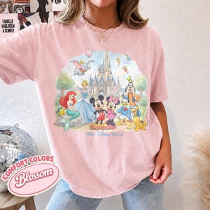 Op de afbeelding: Een lichtroze T-shirt met korte mouwen en een kleurrijke grafische print van Disney-figuren waaronder Mickey Mouse, Minnie Mouse, Donald Duck, Goofy, Pluto, Ari&euml;l, Assepoester en Dumbo voor een kasteel met de tekst Walt Disney World.