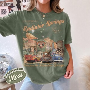 May include: A moss green short-sleeve t-shirt featuring a graphic print of Radiator Springs with text that reads California Est 2012 Radiator Springs and Welcome to Radiator Springs, depicting three cars, a water tower, and desert rock formations.