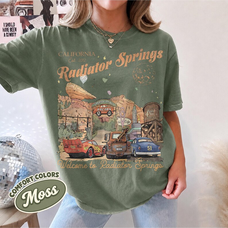 May include: A moss green short-sleeve t-shirt featuring a graphic print of Radiator Springs with text that reads California Est 2012 Radiator Springs and Welcome to Radiator Springs, depicting three cars, a water tower, and desert rock formations.