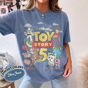 May include: A blue jean colored t-shirt featuring a graphic print of Toy Story characters including Woody, Buzz Lightyear, and others. The text reads Toy Story 5 and You've Got A Friend In Me. The design includes clouds, stars, and various colorful movie figures.