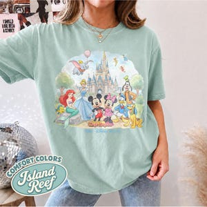 Op de afbeelding: Een lichtgroen t-shirt met korte mouwen en een kleurrijke grafische print van diverse tekenfilmfiguren voor een kasteel. Het ontwerp bevat Dumbo, Ariel, Assepoester, Mickey Mouse, Minnie Mouse, Donald Duck, Goofy en Pluto.