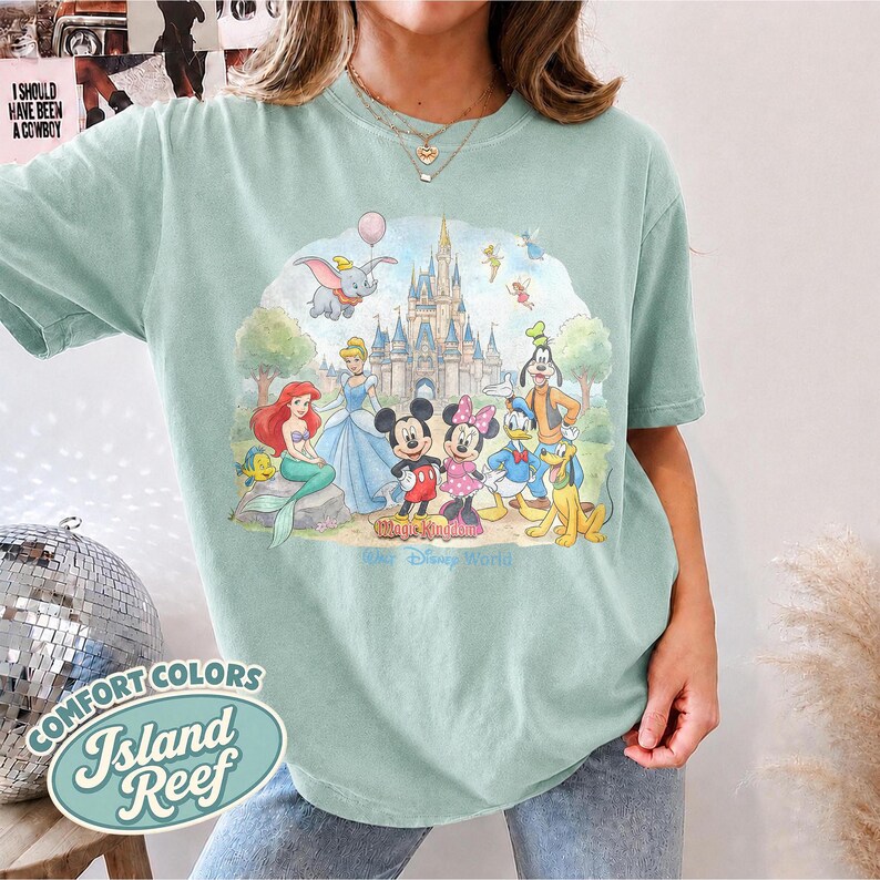 Op de afbeelding: Een lichtgroen t-shirt met korte mouwen en een kleurrijke grafische print van diverse tekenfilmfiguren voor een kasteel. Het ontwerp bevat Dumbo, Ariel, Assepoester, Mickey Mouse, Minnie Mouse, Donald Duck, Goofy en Pluto.