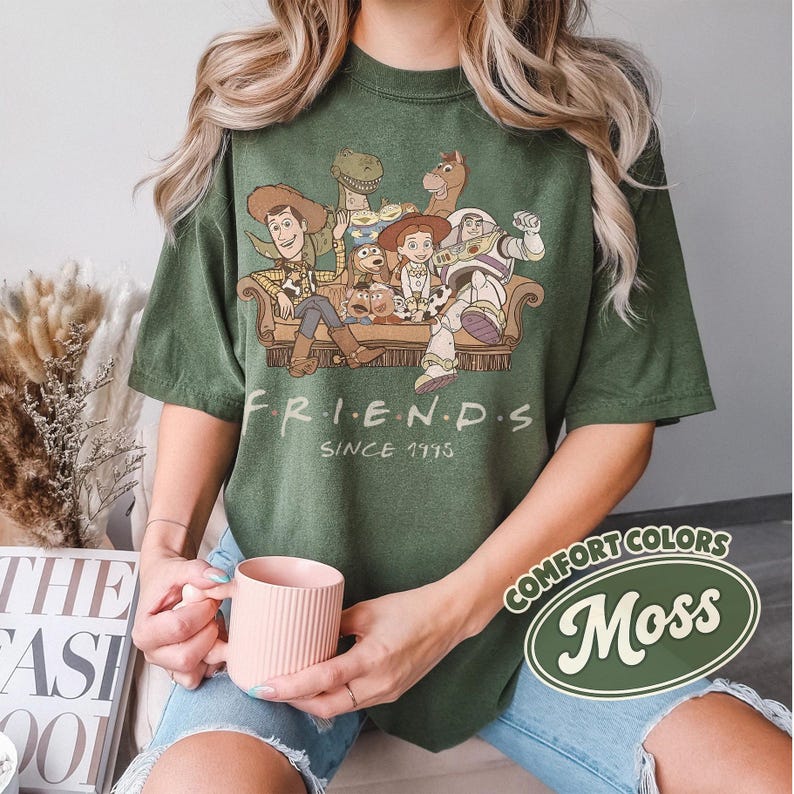 Puede incluir: Camiseta verde con un gr&aacute;fico de personajes de Toy Story en un sof&aacute;, con el texto "FRIENDS" y "SINCE 1995". La camiseta es de la marca Comfort Colors en color "Moss".