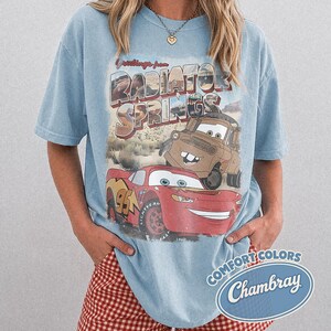 May include: Light blue t-shirt featuring a graphic of Lightning McQueen and Mater from the movie Cars, with the text "Greetings from Radiator Springs." The shirt has a vintage, distressed look and a "Comfort Colors Chambray" logo.