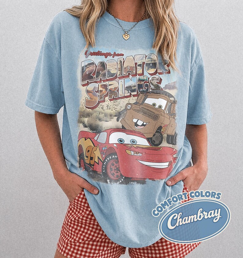 May include: Light blue t-shirt featuring a graphic of Lightning McQueen and Mater from the movie Cars, with the text "Greetings from Radiator Springs." The shirt has a vintage, distressed look and a "Comfort Colors Chambray" logo.