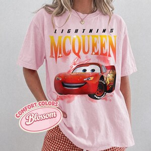 May include: Pink t-shirt featuring a graphic of Lightning McQueen from the Cars movie. The design includes the text "LIGHTNING MCQUEEN" in yellow and orange, and the Comfort Colors Blossom logo.