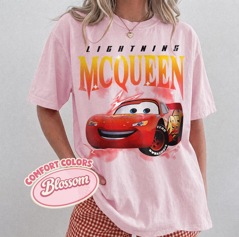 May include: Pink t-shirt featuring a graphic of Lightning McQueen from the Cars movie. The design includes the text "LIGHTNING MCQUEEN" in yellow and orange, and the Comfort Colors Blossom logo.