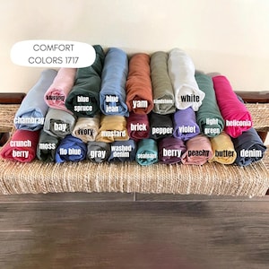 May include: A collection of rolled fabric in various colors, including chambray, blossom, blue spruce, blue jean, yam, sandstone, white, light green, and heliconia. The text "COMFORT COLORS 1717" is displayed at the top. Other colors include crunch berry, moss, bay, ivory, mustard, brick, pepper, violet, peachy, butter, and denim.