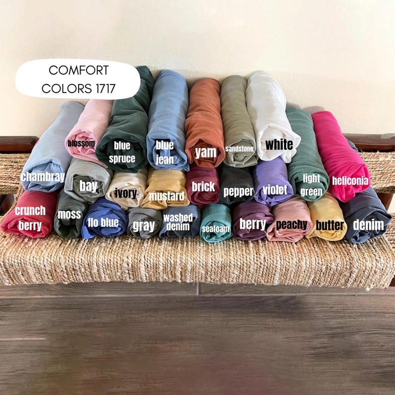 May include: A collection of rolled fabric in various colors, including chambray, blossom, blue spruce, blue jean, yam, sandstone, white, light green, and heliconia. The text "COMFORT COLORS 1717" is displayed at the top. Other colors include crunch berry, moss, bay, ivory, mustard, brick, pepper, violet, peachy, butter, and denim.